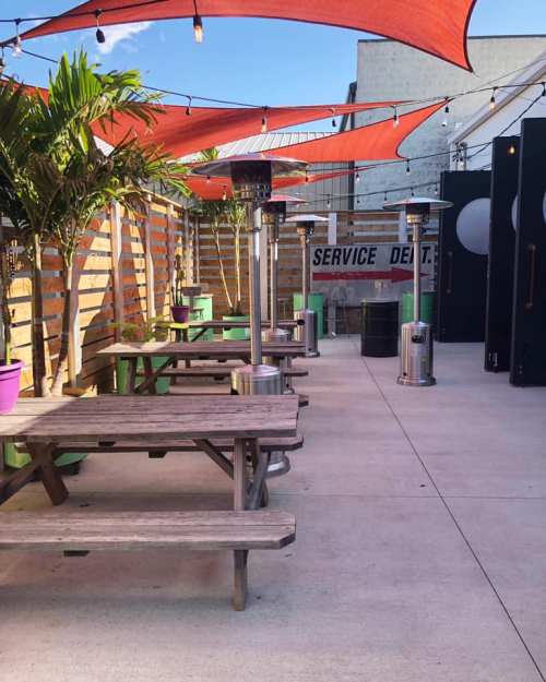 Outdoor seating area with wooden tables, patio heaters, and red canopies, surrounded by greenery and a service sign.
