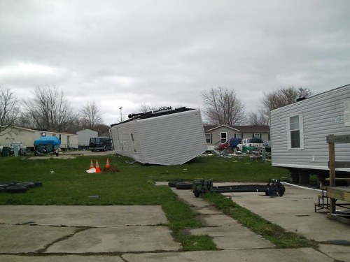 A tilted mobile home in a trailer park, surrounded by debris and other mobile homes under a cloudy sky.