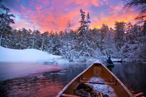 A canoe glides through a snowy landscape at sunset, with vibrant pink and orange skies reflecting on the water.