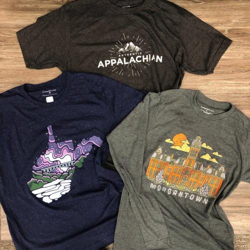 Three graphic t-shirts featuring designs of West Virginia landmarks and "Authentic Appalachian" text on a wooden background.