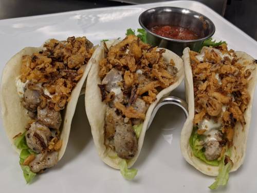 Three tacos filled with meat, lettuce, and crispy onions, served with a side of salsa in a small metal bowl.