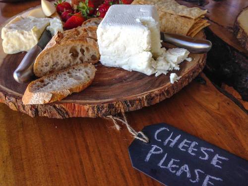 A wooden platter with assorted cheeses, bread, strawberries, and a tag that says "Cheese Please."