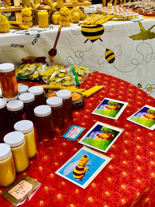 A vibrant market display featuring jars of honey, beeswax candles, and colorful bee-themed artwork on a red tablecloth.