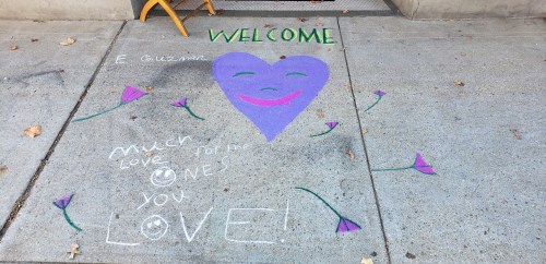 Chalk art on pavement with a purple heart, smiling face, and text: "Welcome E. Guzman. Much love for the ones you love!"