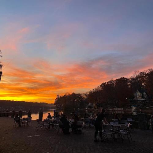 A serene lakeside scene at sunset, with tables set up and people enjoying the view of colorful skies and water.
