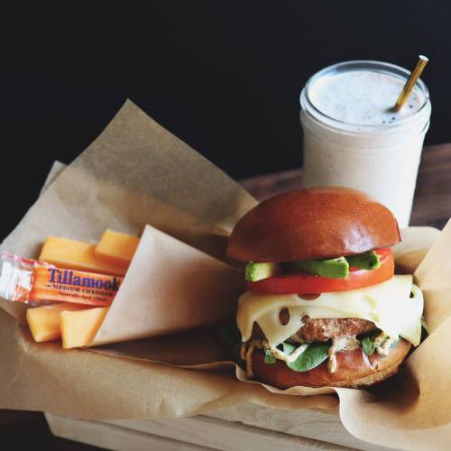 A delicious burger with cheese, lettuce, tomato, and avocado, served with a side of cheese and a milkshake in a jar.