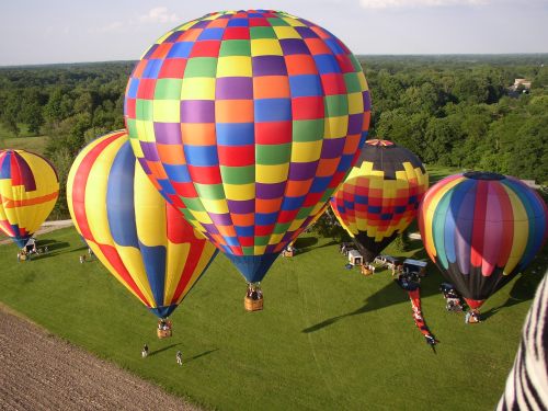 Colorful hot air balloons on a grassy field, with trees in the background and people gathered below.