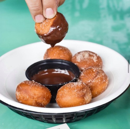 A hand dips a donut hole into a small cup of chocolate sauce, surrounded by more donut holes on a plate.