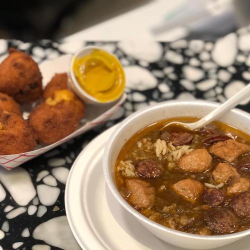A bowl of gumbo with rice and meat, accompanied by cornbread fritters and a side of mustard.