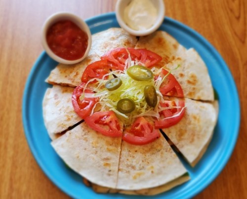 A blue plate with sliced quesadilla topped with lettuce, tomatoes, and jalapeños, served with salsa and sour cream.