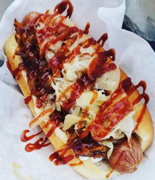 A loaded hot dog topped with sauerkraut, bacon, and drizzled with barbecue sauce, served in a bun.