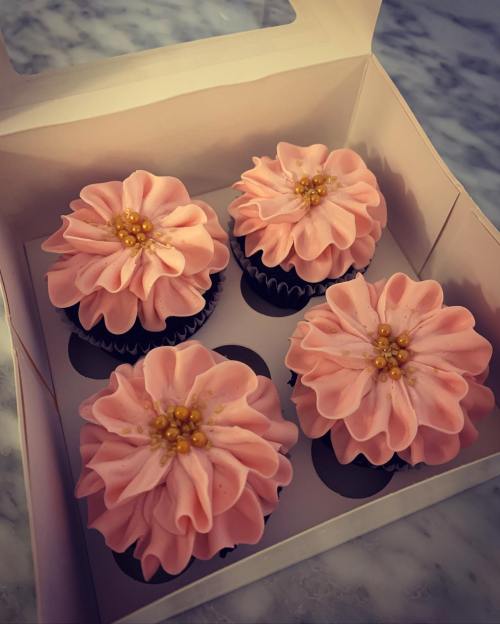 A box of four pink flower-shaped cupcakes with golden sprinkles on a marble surface.