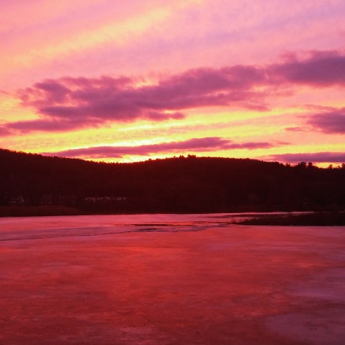 Vibrant sunset over a frozen lake, with colorful clouds reflecting on the icy surface and hills in the background.