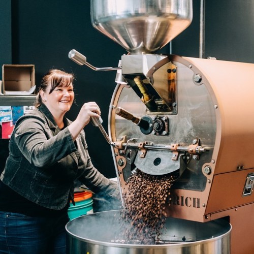 A woman operates a coffee roaster, smiling as freshly roasted beans pour into a container below.