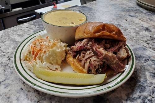 A plate with a pulled pork sandwich, coleslaw, a pickle slice, and a bowl of soup on a marble countertop.
