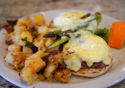 A plate of eggs Benedict with asparagus, served with seasoned potatoes and a slice of orange.