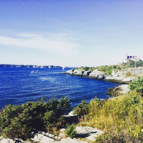 A scenic view of a rocky coastline with blue water, sailboats in the distance, and clear skies.
