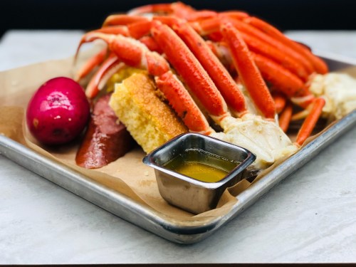 A platter of crab legs, sausage, corn, and a baked potato, served with melted butter on a metal tray.