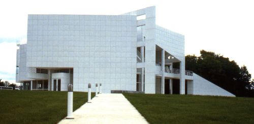 Modern white building with geometric shapes, surrounded by green grass and a pathway leading to its entrance.