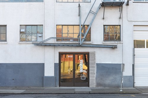 A gray and white building facade with a glass door labeled "408" and a fire escape above. Windows reflect the surroundings.