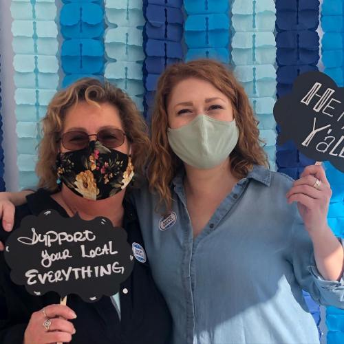 Two women pose together, wearing masks and holding signs that say "Support your local everything" and "Hey y'all!"