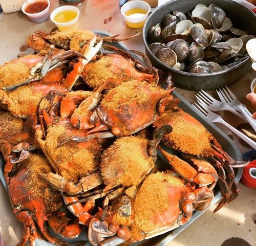 A platter of seasoned crabs with a side of clams, served on a table with utensils and dipping sauces.
