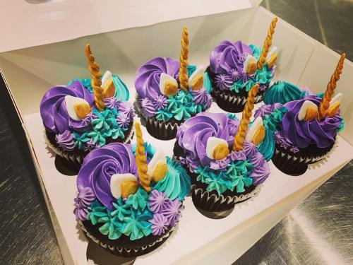 A box of eight colorful unicorn-themed cupcakes with purple and teal frosting, topped with golden horns and decorations.