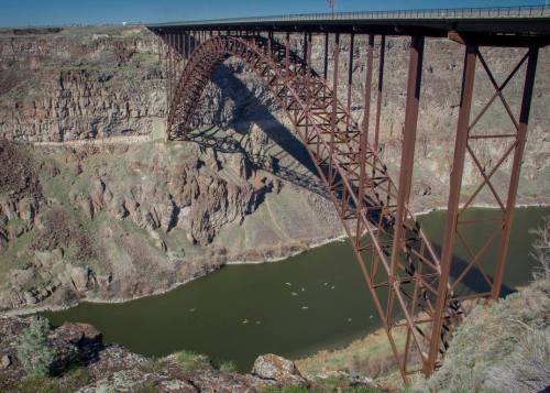 A large metal arch bridge spans a deep canyon over a river, surrounded by rocky terrain and greenery.