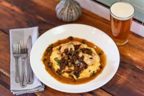 A plate of shrimp and grits topped with mushrooms, served with a glass of beer on a wooden table.