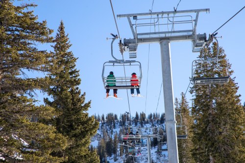 Two skiers on a chairlift, surrounded by snow-covered trees and mountains under a clear blue sky.