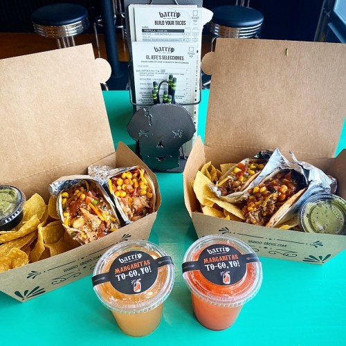 Two takeout boxes with tacos and chips, accompanied by margaritas, on a colorful table. Menu in the background.