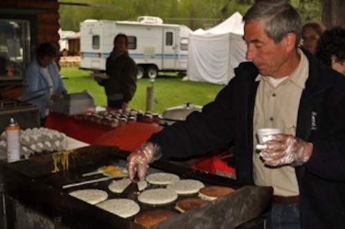 A man cooks pancakes on a griddle at an outdoor event, with people and food stalls in the background.