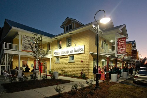 A brightly lit boarding house at dusk, with people gathered outside and decorative signage.