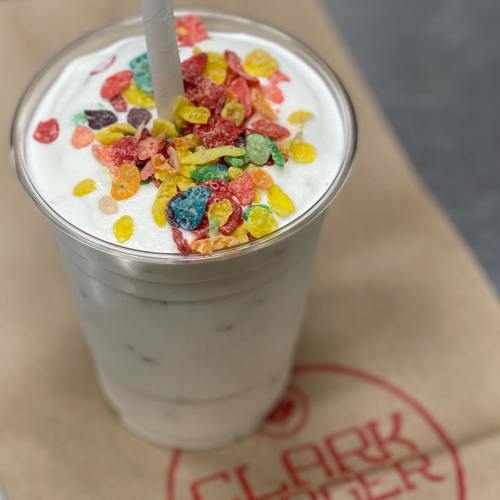 A milkshake topped with colorful cereal pieces in a clear cup on a brown paper surface.