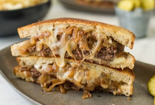 A close-up of a grilled sandwich filled with melted cheese, caramelized onions, and ground beef, served on a plate.