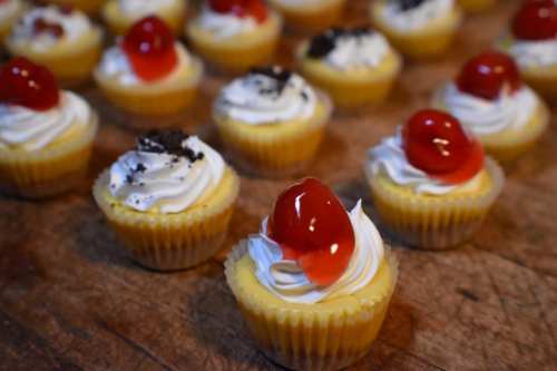 A close-up of mini cheesecakes topped with whipped cream and cherries, arranged on a wooden surface.