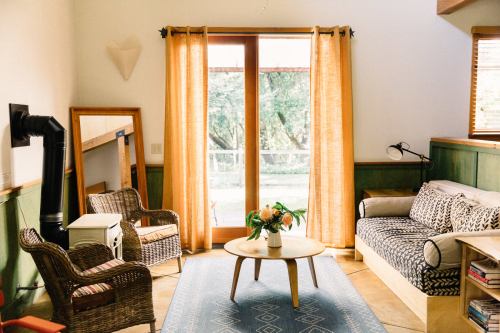Cozy living room with a sofa, armchairs, a coffee table, and large windows with orange curtains overlooking greenery.