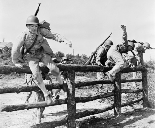 Soldiers in uniform jumping over a wooden fence during training exercises, with rifles slung over their shoulders.