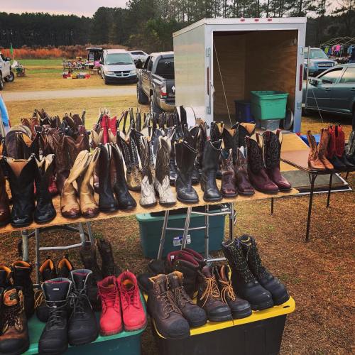 A variety of boots displayed on tables at an outdoor market, with trucks and a trailer in the background.