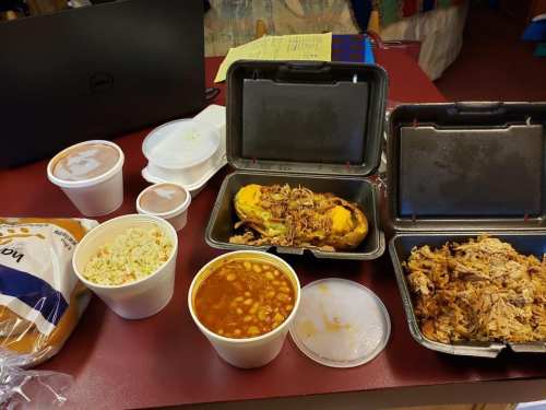 A table with takeout containers of pulled pork, coleslaw, baked beans, and bread rolls, alongside a laptop.
