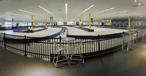 Indoor go-kart track with a winding course, surrounded by barriers and seating areas. Brightly lit and spacious.
