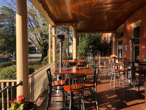 A sunny outdoor patio with wooden tables and chairs, surrounded by greenery and warm lighting.