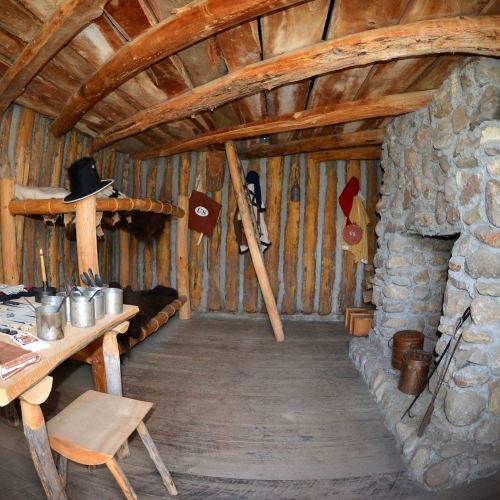 Interior of a rustic wooden cabin with a stone fireplace, wooden beams, bunk beds, and simple furnishings.