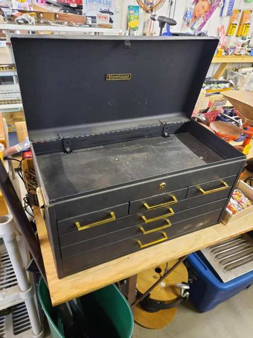An open black tool chest with gold handles, displaying empty drawers and a spacious interior on a workbench.