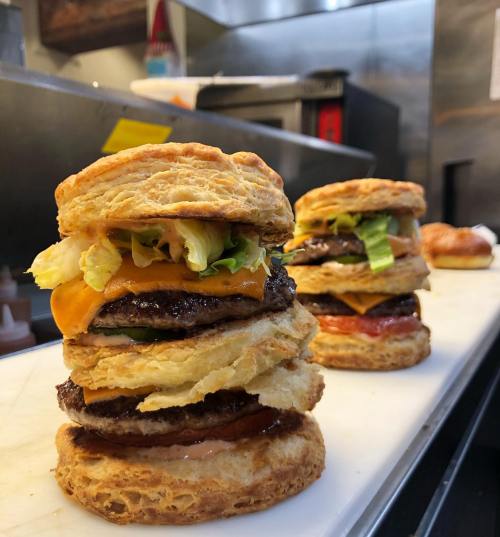 Three stacked burgers with flaky biscuit layers, lettuce, cheese, and various meats, displayed in a kitchen setting.