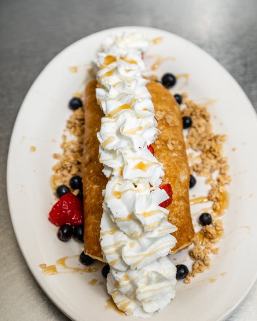 A golden crepe filled with strawberries and blueberries, topped with whipped cream and drizzled with syrup on a plate.