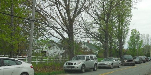 A tree-lined road with parked cars on one side and a white house in the background, under a cloudy sky.