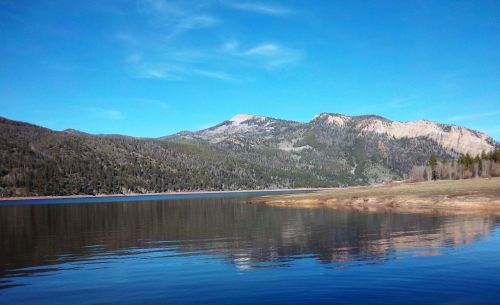 A serene lake reflects mountains and a clear blue sky, surrounded by lush greenery and a sandy shore.