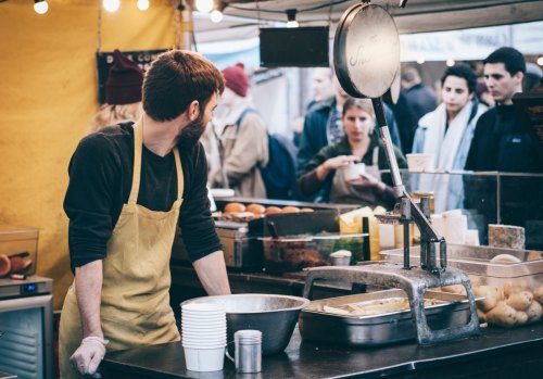 A vendor in an apron stands at a market stall, preparing food while customers gather in the background.