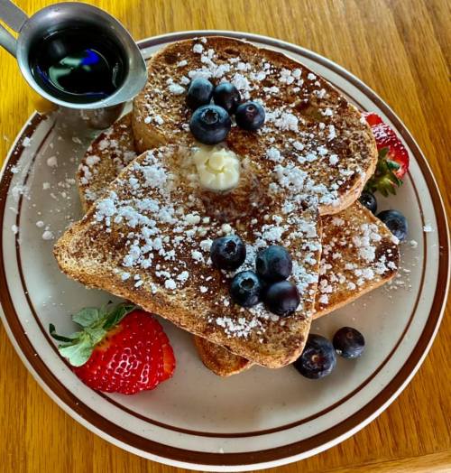 A plate of French toast topped with powdered sugar, blueberries, and strawberries, with a small cup of syrup on the side.